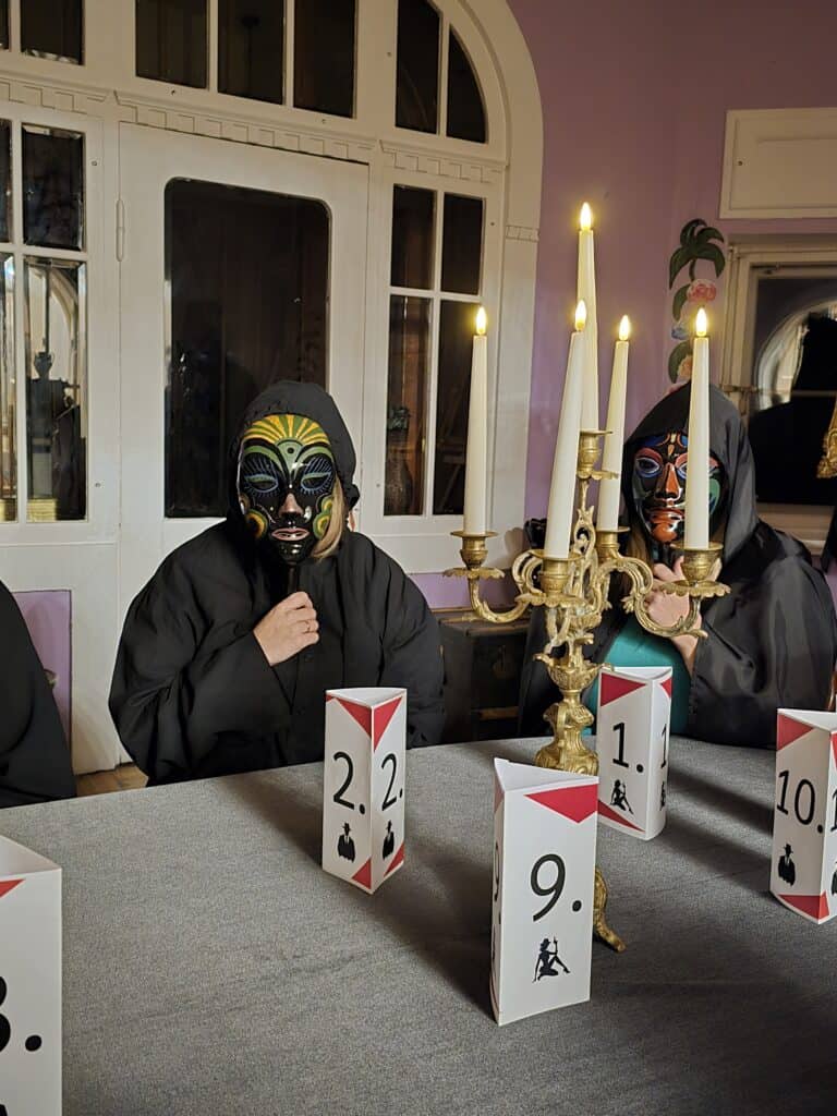 Divi cilvēki ar maskām spēlē mafijas un parlamenta spēli pie galda ar skaitļiem un svečturiem.