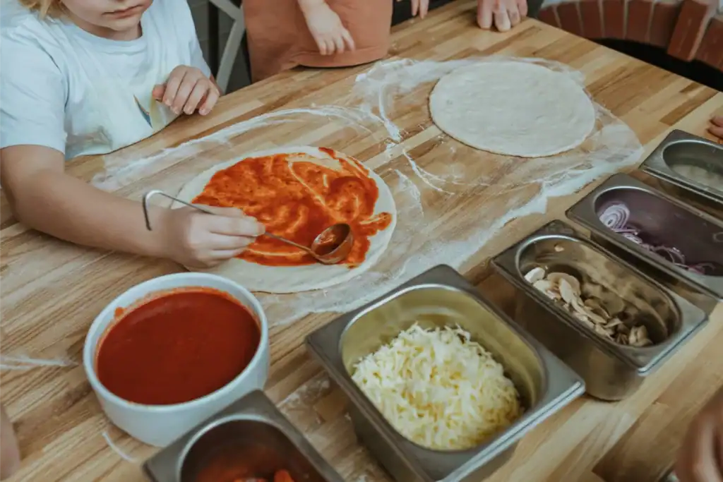Vaikai gamina picos tešlą, naudodami ingredientus ir įrankius.