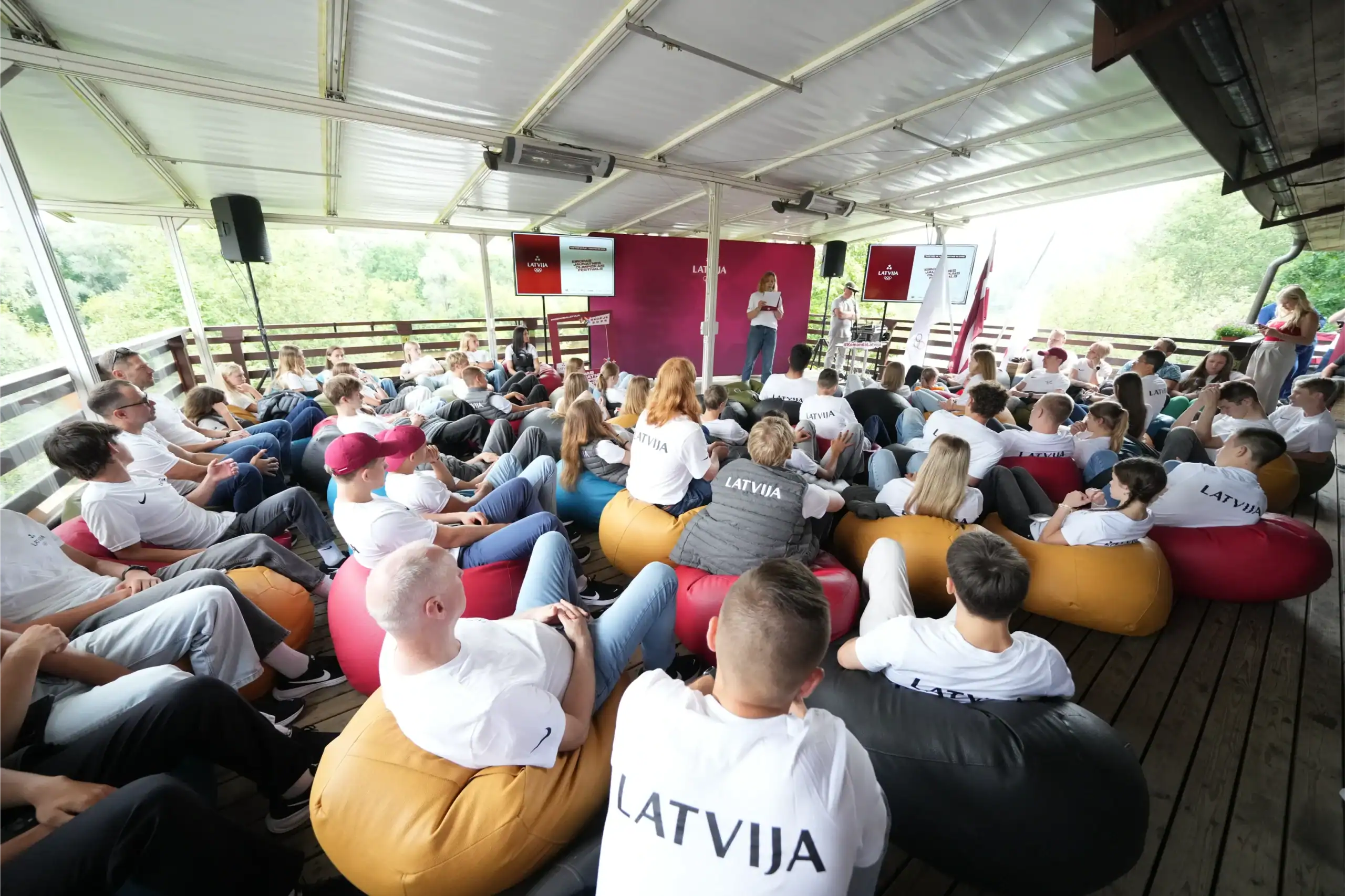 Grupės nariai sėdi ir klausosi pasirodymo po atviru dangumi, kur yra vitrina ir scena.
