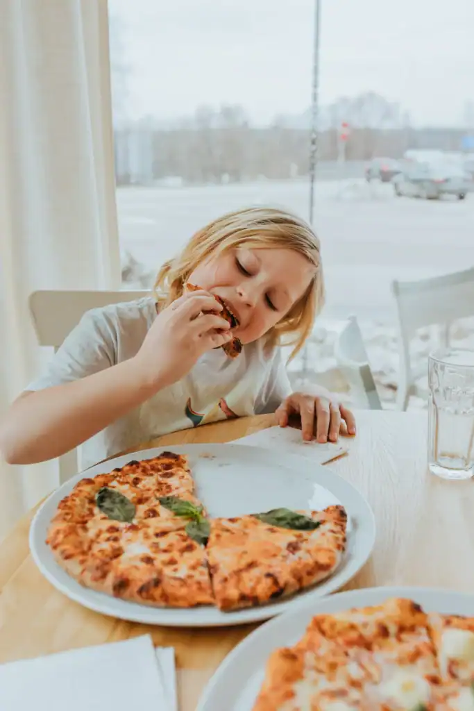 Jauns bērns ar blondiem matiem ēd picu pie galda, ēdināšanas vietā ar lielu logu un pilsētas.