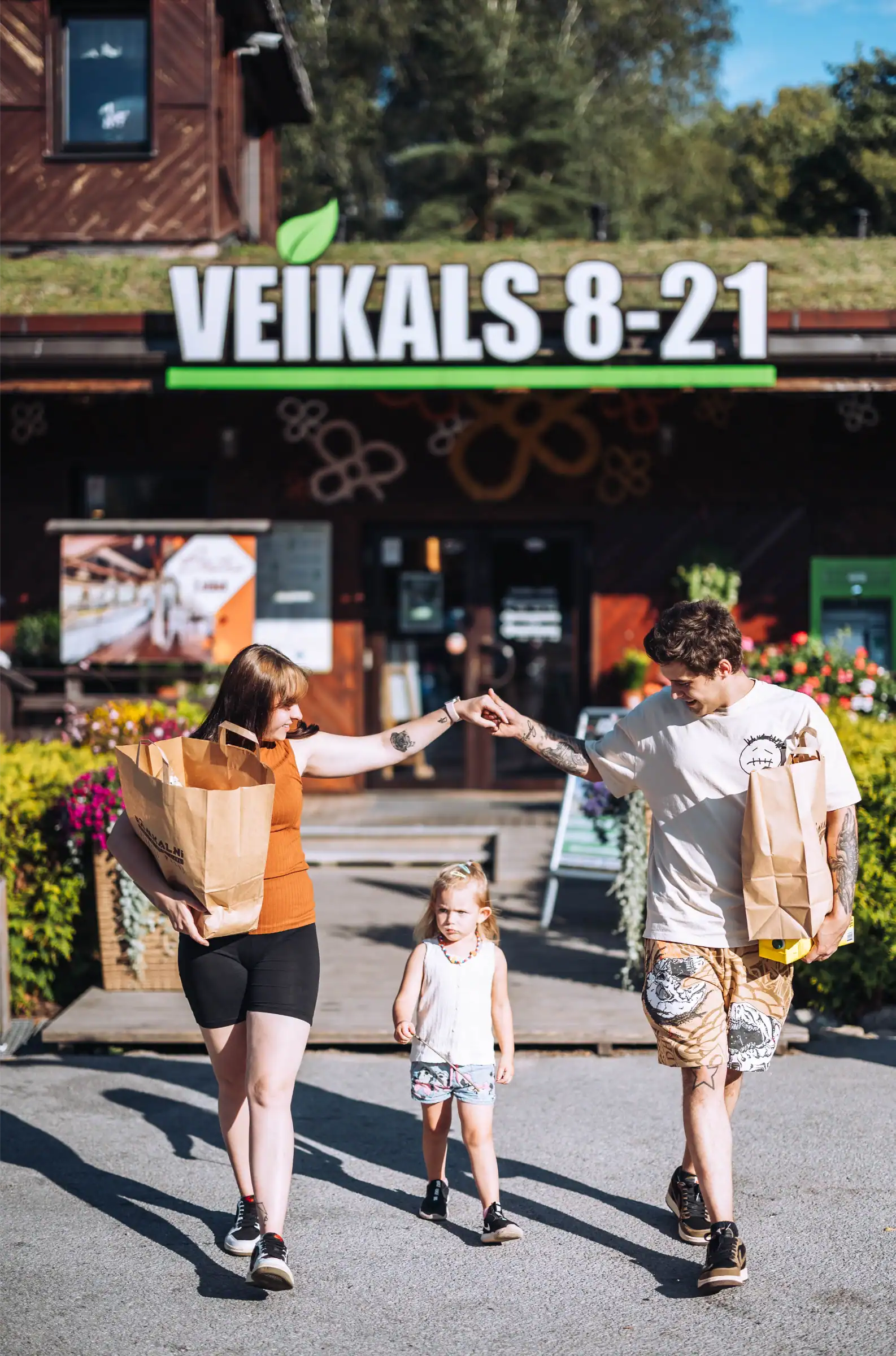 Veselīga ģimene ar bērnu, iepērkoties veikalā Veikals 8-21, ārpusē, saulainā dienā.
